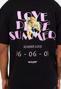 Camiseta de algodón negra con texto grande en rosa "AMOR PAZ VERANO" junto a figuras angelicales, y texto adicional "AMOR VERANO 06-06-08."