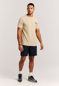 Beige katoenen T-shirt met een afbeelding op de borst, gecombineerd met donkerblauwe shorts en zwart/grijze sneakers met een golvend patroon. Witte enkelsokken zichtbaar.