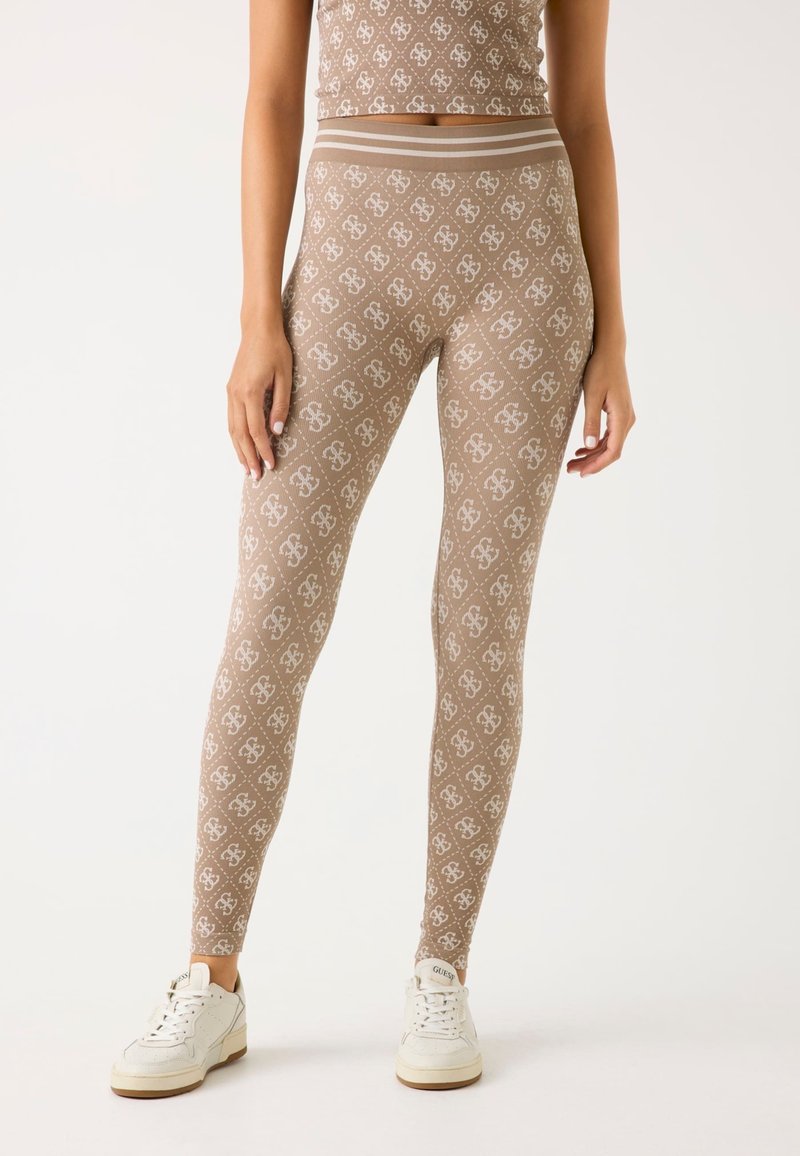 Modella che indossa leggings beige e un crop top abbinato con un motivo a logo bianco ripetuto, abbinato a sneakers bianche su uno sfondo neutro.