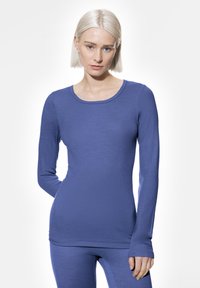 Femme jeune avec les cheveux platine blonds courts portant un t-shirt ajusté à manches longues bleu et un pantalon assorti, se tenant devant un fond blanc.