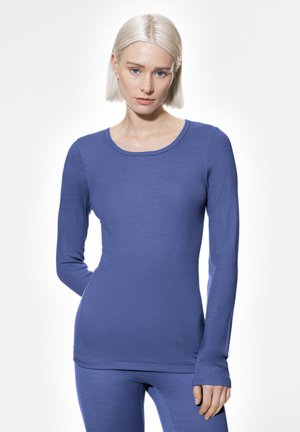 Junge Frau mit kurzem platinblondem Haar, die ein enganliegendes, langärmliges blaues Shirt und passende Hosen trägt, steht vor einem weißen Hintergrund.