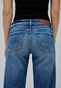 Vaqueros de mezclilla azul medio con un ajuste relajado. Las características incluyen bolsillos traseros dobles, textura desgastada y una etiqueta de cuero marrón con "Salsa Jeans."