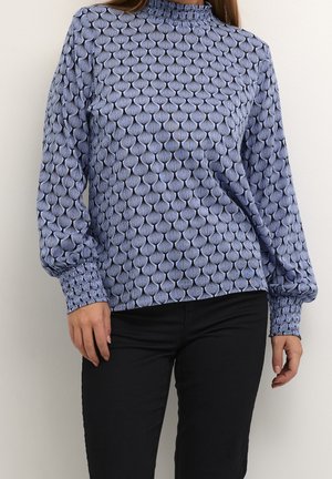 Kvinde iført en langærmet mønstret bluse med høj hals og sorte bukser, stående foran en ensfarvet lys væg.