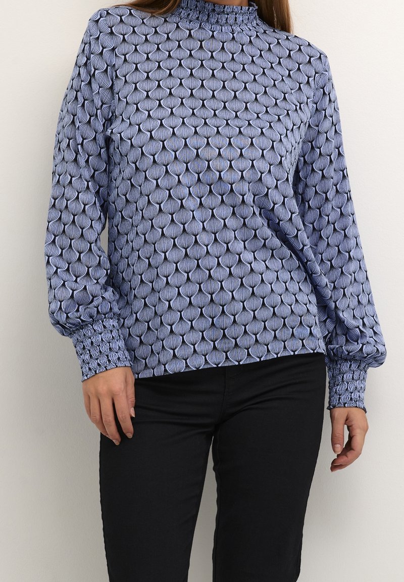 Femme portant un chemisier à manches longues à motifs avec un col montant et un pantalon noir, debout contre un mur clair uni.