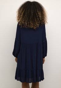 Navyblå knälång klänning med långa ballongsärmar och volangkant. Tillverkad av lätt material med en lös, svävande siluett.