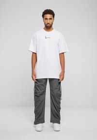 T-shirt blanche en coton avec impression de signature noire, associée à un pantalon cargo gris avec des coutures latérales froncées et des baskets blanches.