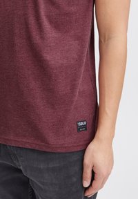 Burgunderfarbenes T-Shirt mit kurzen Ärmeln aus weichem Stoff, mit einem Seitenschild mit Logo. Getragen mit dunkelgrauen Hosen.