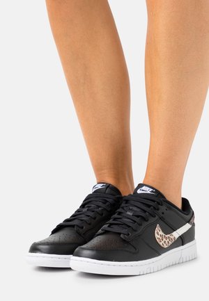 Zwarte Nike sneakers met witte zolen en een luipaardprint swoosh, gedragen zonder sokken tegen een effen witte achtergrond.