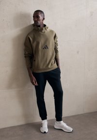 Felpa con cappuccio verde oliva con dettagli neri e tasca a marsupio, abbinata a pantaloni jogger neri a gamba affusolata e scarpe atletiche bianche.