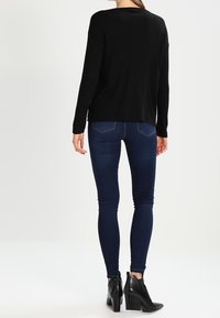 Maglione nero a maniche lunghe in maglia abbinato a jeans skinny blu scuro e stivaletti con tacco neri, visto da dietro.