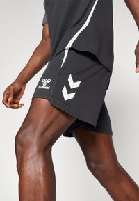 Persona atlética vistiendo pantalones cortos negros de Hummel y una camiseta a juego, capturada en plena zancada con piernas musculosas visibles y movimiento de brazos.