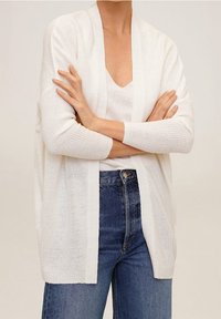 Bílý pletený cardigan s otevřeným velikostí a dlouhými rukávy. Nosený přes odpovídající bílý top a modré džíny s vysokým pasem, které mají uvolněný střih.