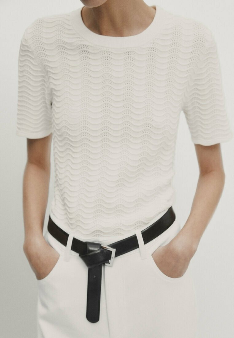 Personne portant un haut blanc texturé à manches courtes avec des motifs en vagues et un pantalon blanc avec une ceinture noire nouée à la taille.