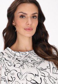 Blusa bianca con motivi a linee astratte nere, scollatura rotonda e tessuto morbido. Il modello indossa orecchini a cerchio dorati e ha capelli scuri e mossi.