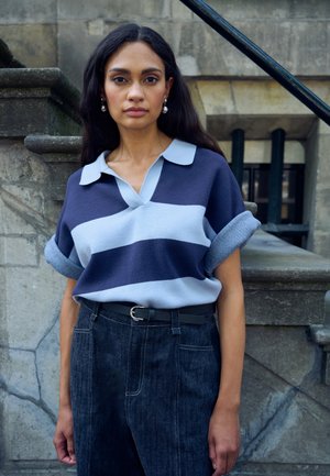 Marineblauw en lichtblauw gestreept poloshirt met kraag, gecombineerd met hooggetailleerde donkere denim broek en een zwarte riem met een zilveren gesp.