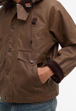 Mężczyzna ubrany w brązową woskowaną bawełnianą kurtkę z kieszeniami na klapę zapinanymi na guziki, kołnierzem i mankietami z sztruksu, z ręką w kieszeni oraz widocznymi metkami Levi's i Barbour.