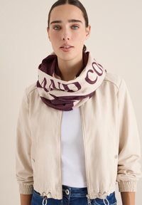 Sciarpa circolare in crema e bordeaux, con un audace design testuale. Il modello indossa una leggera giacca e una camicia bianca, insieme a dei jeans. Texture liscia.