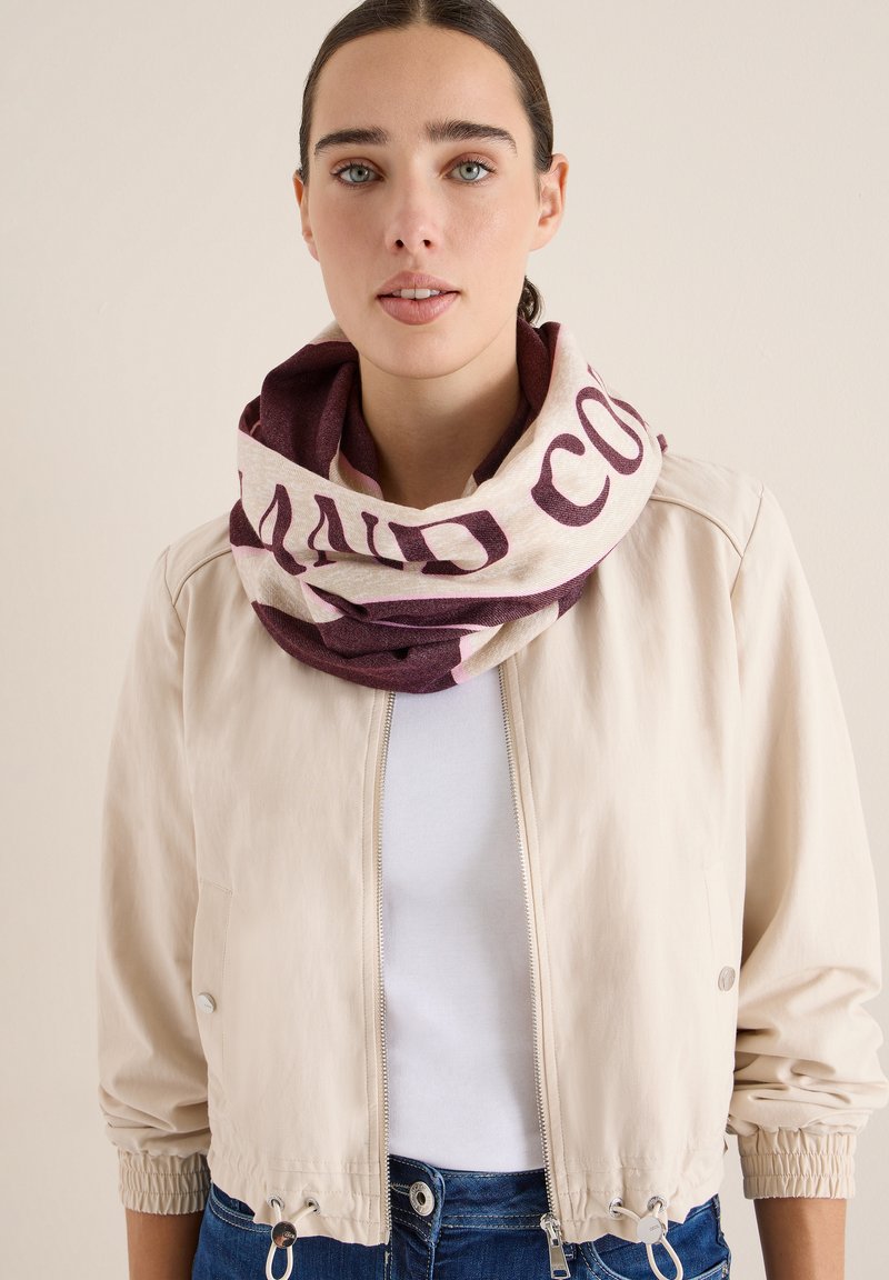 Sciarpa circolare in crema e bordeaux, con un audace design testuale. Il modello indossa una leggera giacca e una camicia bianca, insieme a dei jeans. Texture liscia.