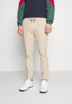 Man wearing beige drawstring pants, white sneakers, and a navy sweatshirt with red and green sleeves standing against a plain background.