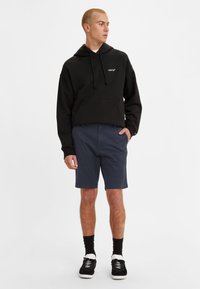 Hanorac negru cu glugă, dotat cu un buzunar tip pouch, asortat cu pantaloni scurți bleumarin. Modelul poartă adidași negri cu talpă albă și șosete negre.