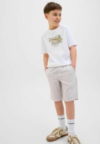 Vit t-shirt med kort ärm och grafiskt tryck, ljusgrå shorts, beige sneakers och vita strumpor med svart text, stående mot en neutral bakgrund.