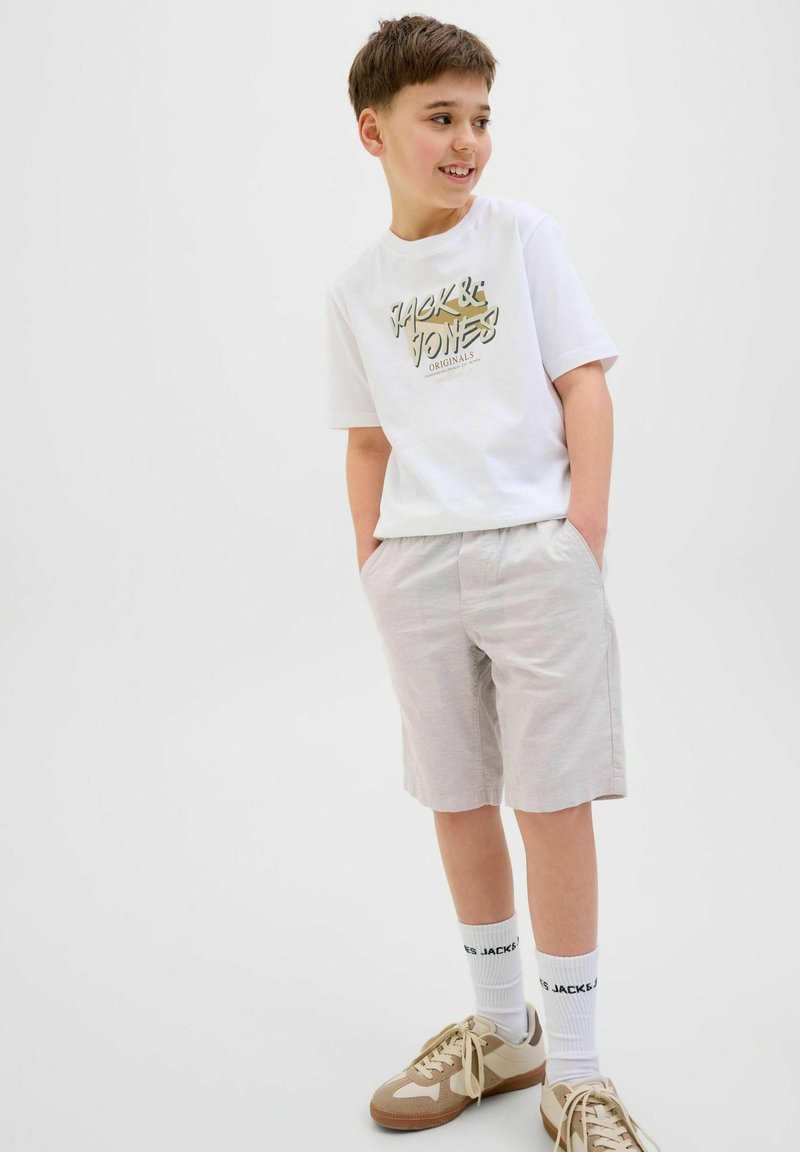 Vit t-shirt med kort ärm och grafiskt tryck, ljusgrå shorts, beige sneakers och vita strumpor med svart text, stående mot en neutral bakgrund.