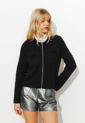 Femme blonde portant une veste noire zippée sur un chemisier blanc à volants et un short argenté métallisé, posant devant un fond blanc.