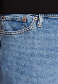 Světle modré džíny mají klasický pětivreckový design, měděné nýty a kontrastní oranžové švy. Texturovaná tkanina s vybledlým odstínem.