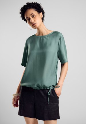 Hellgrünes Satin-Oberteil mit Rundhalsausschnitt, kurzen Ärmeln und gerafftem Saum, der an der Seite gebunden ist. Kombiniert mit schwarzen Shorts. Glatte Textur.