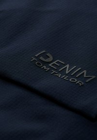 Niebieski materiał z teksturowanym wzorem w kratkę, z wypukłym czarnym logo, które brzmi "DENIM TOM TAILOR". Gładka powierzchnia, lekko błyszczące wykończenie.