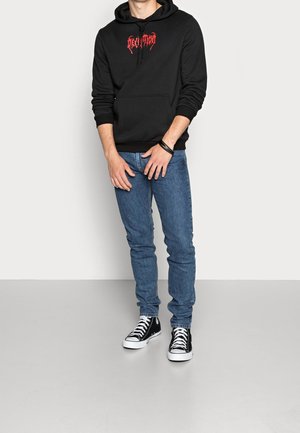 Mężczyzna w czarnej bluzie z kapturem z czerwonym napisem, niebieskich dżinsach, czarnych wysokich trampkach i czarnej opasce na nadgarstek, stojący na białym tle.