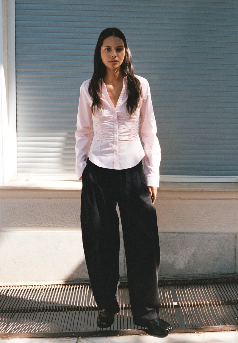Femme aux longs cheveux foncés porte un chemisier rose clair froncé et un pantalon noir ample, debout à l'extérieur devant un mur beige avec un volet fermé.
