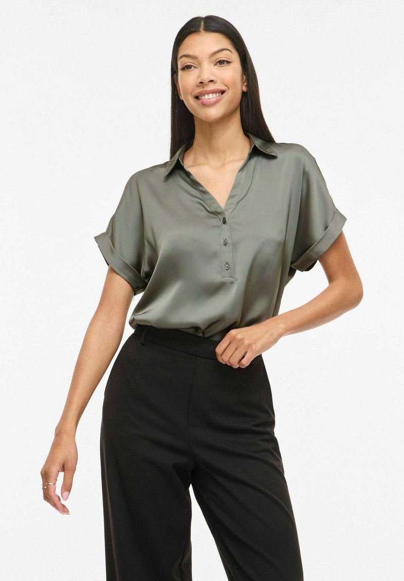 Femme souriante portant un chemisier en satin vert olive à manches courtes avec des boutons et un pantalon noir taille haute, posant devant un fond blanc.