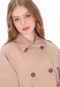 Beige trenchcoat met grote gouden knopen in de vorm van een anker, voorzien van een brede kraag en een gladde, glanzende textuur.