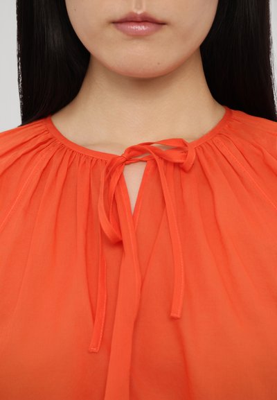 Leuchtend orangefarbene Bluse mit runder Ausschnitt. Verfügt über Raffung an den Schultern und einen Frontknotenverschluss. Hergestellt aus einem leichten Stoff.