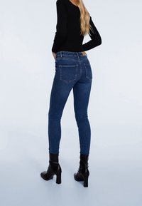 Jeans ajustados de mezclilla azul con cintura alta, dos bolsillos traseros y una etiqueta de cuero marrón en la cinturilla, combinados con botas de tacón negras.