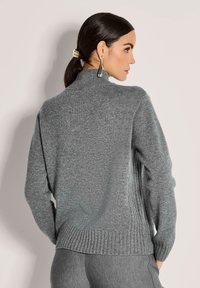 Frau in grauem Strickpullover und passender Hose, die seitlich steht, mit Haaren in einem tiefen Pferdeschwanz, der mit einer goldenen Haarspange fixiert ist, und trägt silberne Ohrringe.