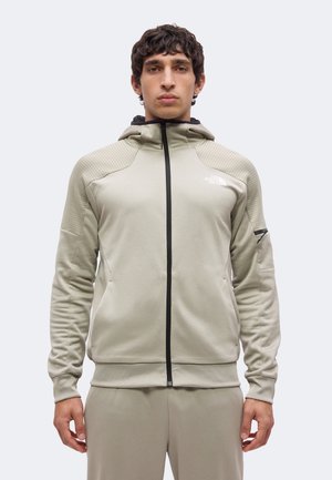 Joven con cabello rizado oscuro que lleva una sudadera con capucha beige clara con cremallera y pantalones a juego sobre un fondo liso y claro.
