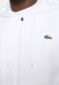 Witte hoodie met ritssluiting, gemaakt van zachte stof. Voorzien van een klein groen Lacoste krokodillenlogo op de linkerborst en platte trekkoorden.