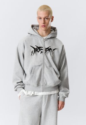 Ung mand med kort blondt hår iført en grå lynlåshoodie med sort grafisk tekst og matchende grå joggingbukser, hænderne i lommerne.