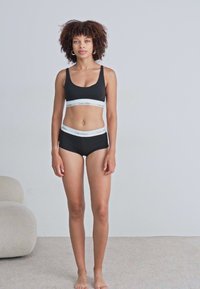 Reggiseno sportivo in cotone nero e mutande abbinate con il logo bianco "Calvin Klein" sulla cintura. Il modello è in piedi a piedi nudi su un tappeto chiaro.