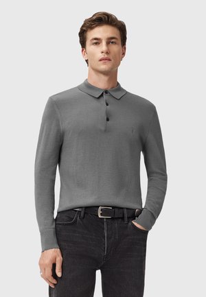 Mand iført grå langærmet polo med sorte knapper, sort bælte og mørke jeans, stående med en hånd i lommen foran en ensfarvet baggrund.