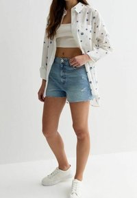 Donna che indossa un crop top bianco, una camicia bianca oversize con motivi a conchiglia blu, pantaloni shorts di denim e sneakers bianche, in piedi contro uno sfondo semplice.