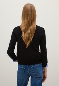 Suéter negro de manga larga, usado sobre una camisa azul clara, combinado con jeans de mezclilla azules que tienen bolsillos traseros. El cabello es largo y ondulado.