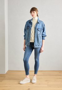Giacca di jeans blu chiaro sopra un maglione a treccia color crema, abbinata a jeans skinny blu scuro e sneaker beige. Design semplice con texture sottili.