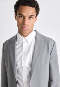 Blazer gris con solapas en muesca sobre una camisa blanca de botones. Presenta cierre de un solo botón, tela suave y un corte entallado.