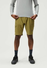 Olivgrüne Cargo-Shorts mit zwei seitlichen Taschen, kombiniert mit einem hellgrauen Langarmshirt und schwarzen Schuhen. Texturierte navyfarbene Socken mit Muster sind sichtbar.