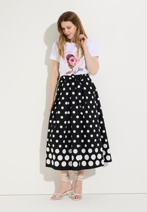 Frau trägt weißes Grafik-T-Shirt und schwarzen High-Waist-Midirock mit großen weißen Punkten, steht vor weißem Hintergrund.