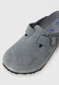 Cloguri gri din suede, cu talpă neagră și curea reglabilă, văzute dintr-un unghi care evidențiază detaliile de sus și din față.