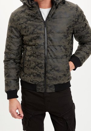 Homme portant une doudoune camouflage numérique verte et noire avec zip devant et poignets et ourlet côtelés noirs, associée à un pantalon cargo noir.